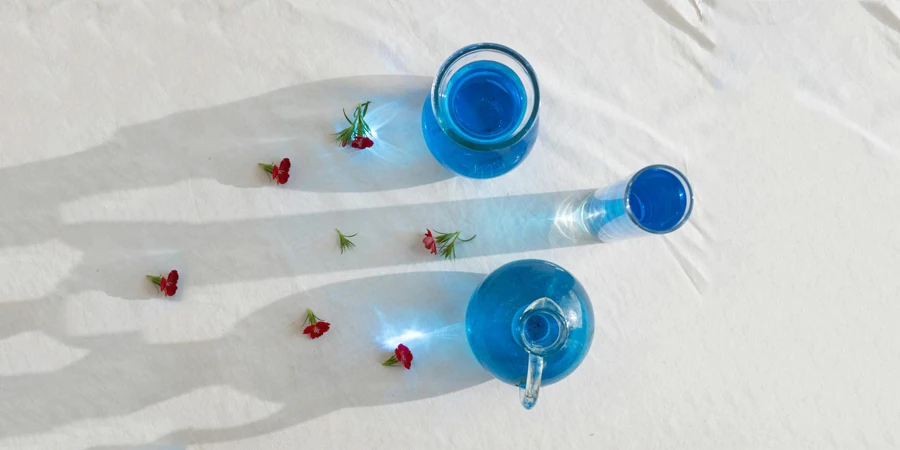 Top view of composed blossoms and various vases and glass with blue aqua on white fabric in sunlight