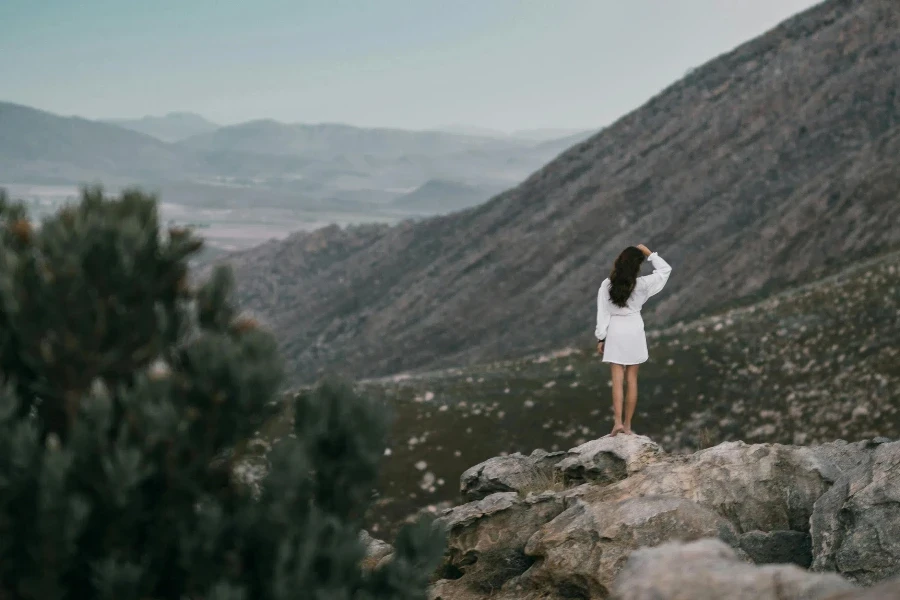 Woman in White Long Sleeves and Skirt Standing on Rocks Near Mountains