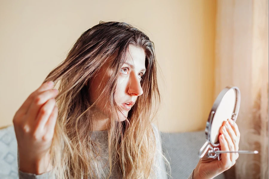 Young annoyed woman is upset about dirty oily and greasy hair looking in mirror at home