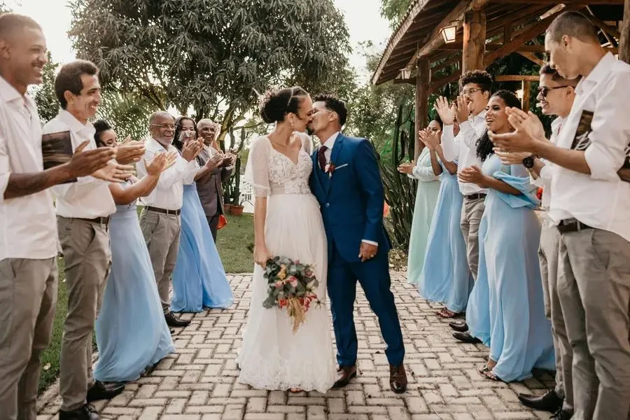 Young bride with flower bouquet kissing black husband between cheerful clapping guests on tiled walkway during festive event by Jonathan Borba