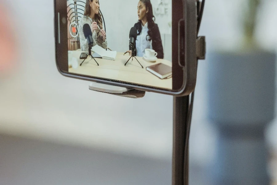 Young diverse women sitting at table with microphones and gadgets and talking while recording podcast on smartphone placed on tripod in studio by George Milton