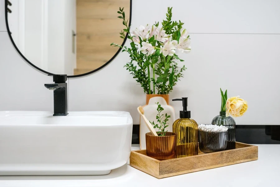 detail in bathroom interior, modern washbasin with faucet, dispenser bottle with soap, toothbrush in glass and cotton ears sticks on wooden tray near vase with flowers