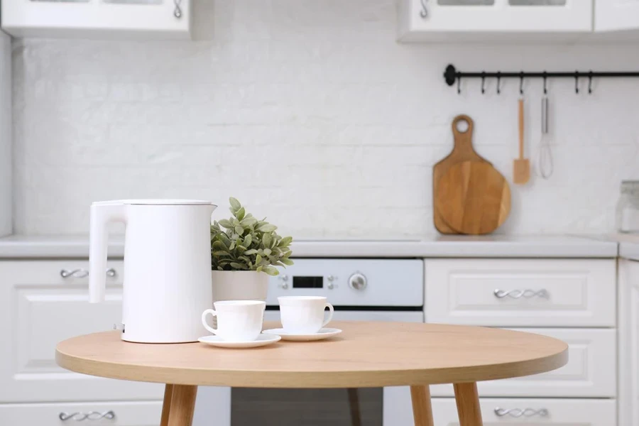 two cups of tea and white electric kettle, green flower in pot on wooden round table in white cozy kitchen. 