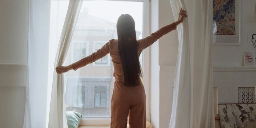 A Back View of a Woman Standing Near the Window while Opening Curtains