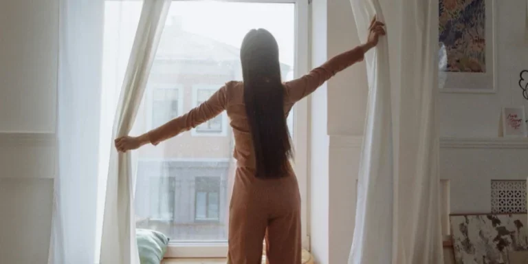 A Back View of a Woman Standing Near the Window while Opening Curtains