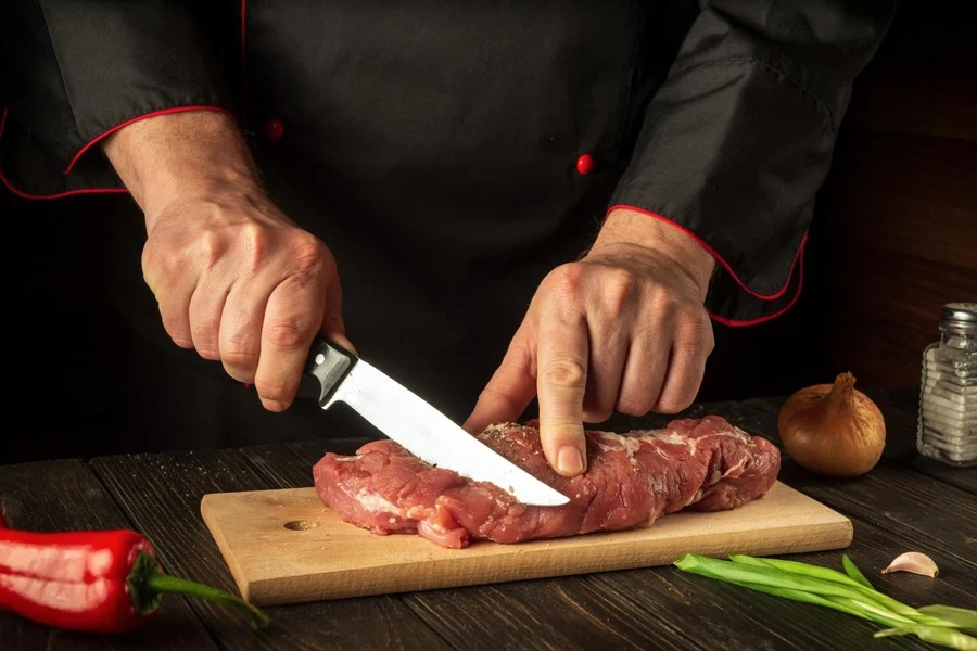 A cook cuts raw veal on a cutting board with a knife before cooking