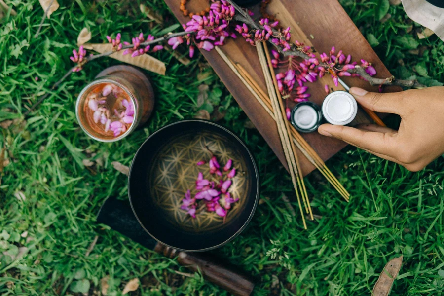 A hand holding a small container over a bowl with pink flowers. Nearby are a glass jar, incense sticks, a wooden tray, and additional pink blossoms spread on grass