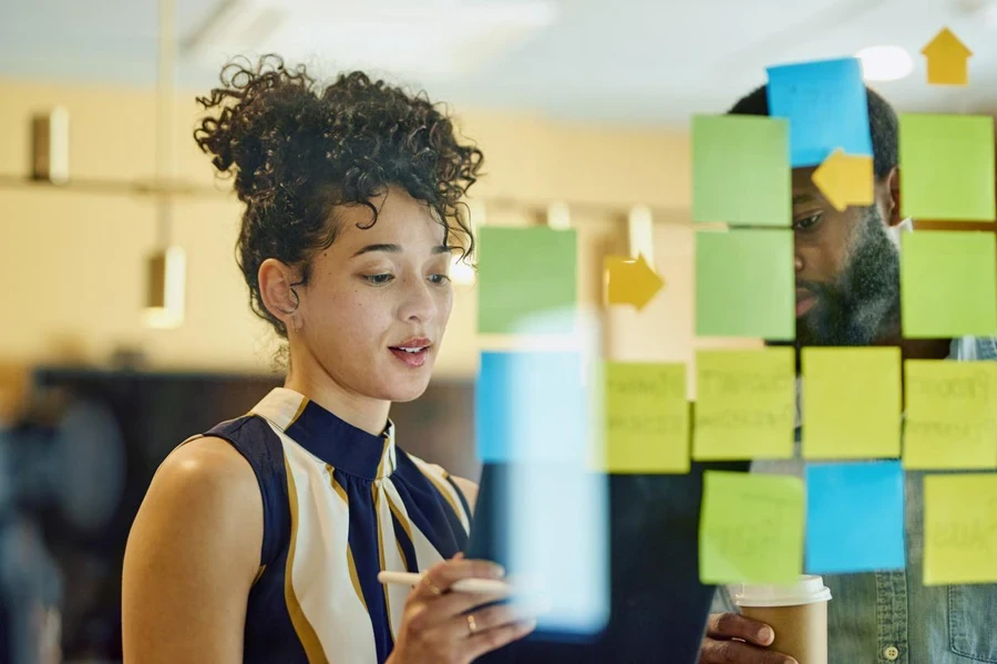 A man and a woman are standing in front of a glass wall writing on sticky notes.