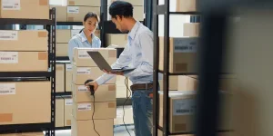 Asian business partner man hold laptop with scanner machine girl wear formal shirt hold tablet discuss inventory online stock data for delivery customer at warehouse