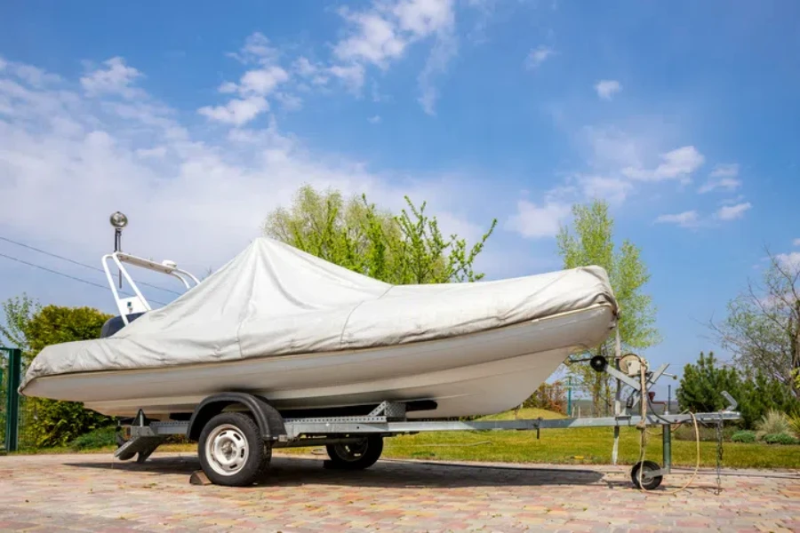 Big modern inflatable motorboat ship covered with grey or white protection tarp standing on steel semi trailer at home backyard on bright sunny day with blue sky on background