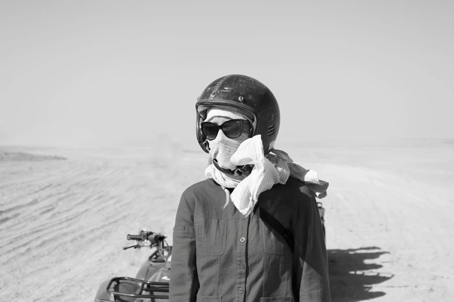 Black and white anonymous biker in helmet covering face with scarf and sunglasses standing near motorbike on sandy terrain 