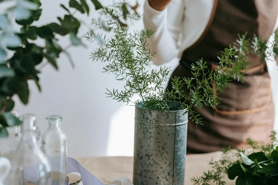 Crop anonymous woman in apron against desk with evergreen plant sprigs in vase in light workspace