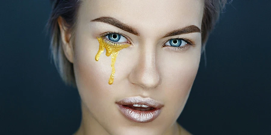 Female model with gold paint of artistic makeup under eye and metal lipstick on lips looking at camera