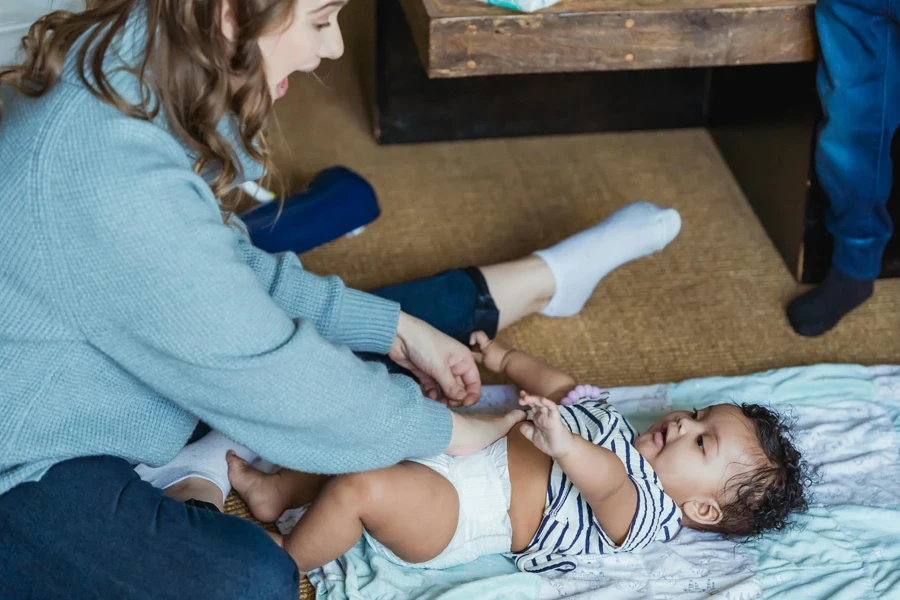 From above excited young mother changing diaper and playing with adorable ethnic baby lying on linen on floor