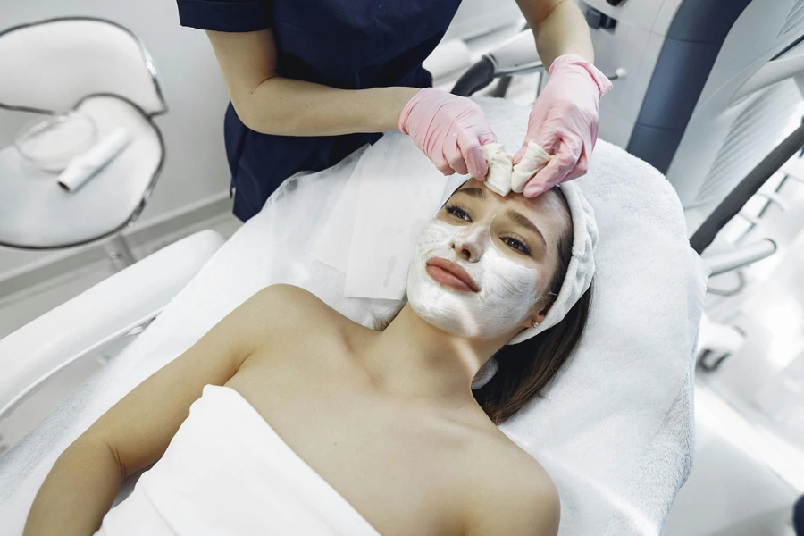 From above of anonymous specialist providing face cleaning for attractive female in beauty salon