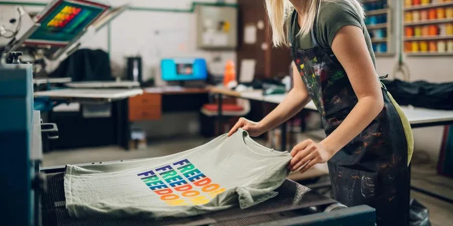 Happy print shop worker putting screen printed t-shirt in drying machine stock photo