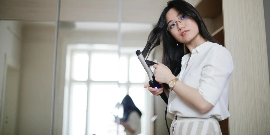 Low angle of stylish Asian female curling hair with iron and looking away in morning at home