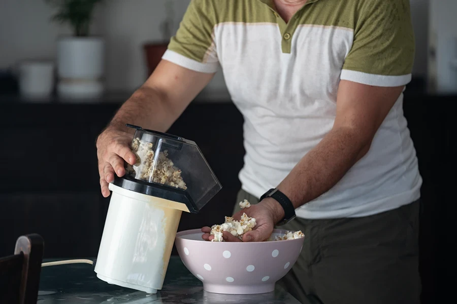 Photo of mature adult man standing in domestic kitchen and cooking popcorns with hot air popper