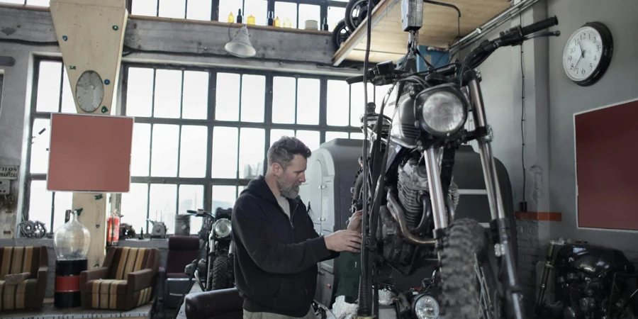 Side view of bearded adult mechanic in casual uniform standing near motorcycle and working with engine in workshop