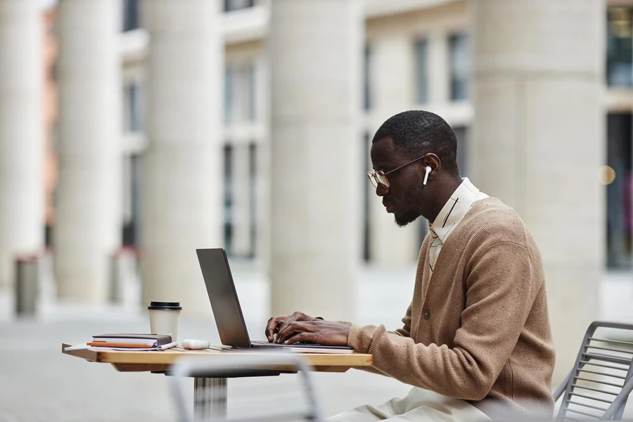 Side view of young African American businessman in earphones and casualwear sitting by table in front of laptop and networking in street cafe