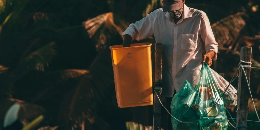 Unrecognizable black male in casual wear and cap collecting trash from street garbage bins in tropical country by Thom Gonzalez