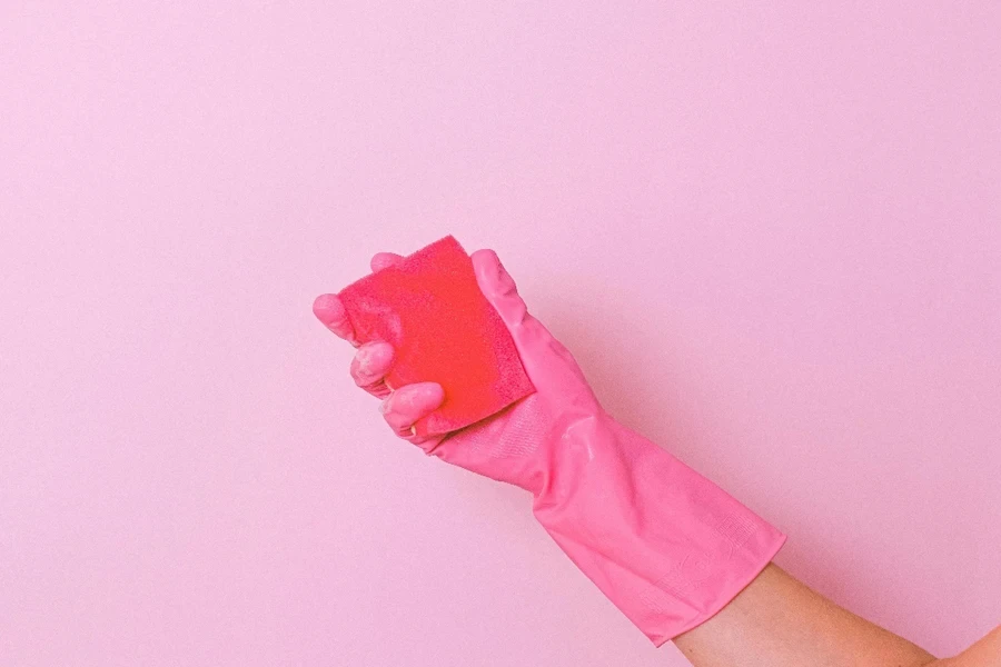 Unrecognizable housekeeper wearing latex glove standing on pink background with sponge in hand for cleaning dirt surface in bright studio