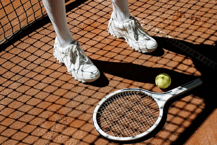 Person Wearing White Sneakers Standing Beside a Tennis Racket