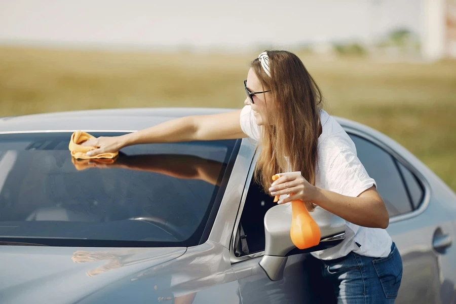 Smiling young woman wiping automobile in countryside during car travel