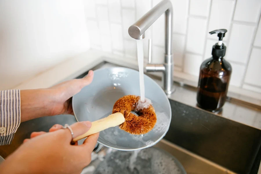 Unrecognizable female with brush washing plate at sink with pouring water and detergent while standing in light kitchen at home