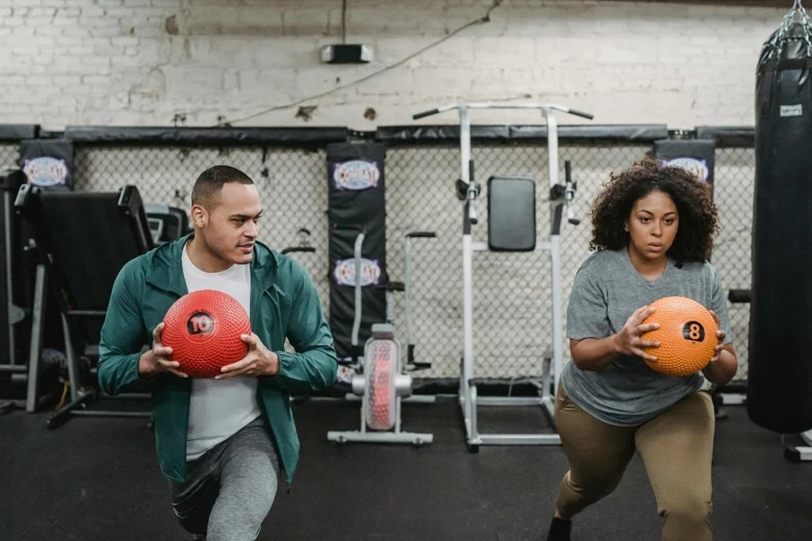 Young muscular ethnic male instructor in activewear doing lunges exercise with ball near plus size black woman during training in sport club