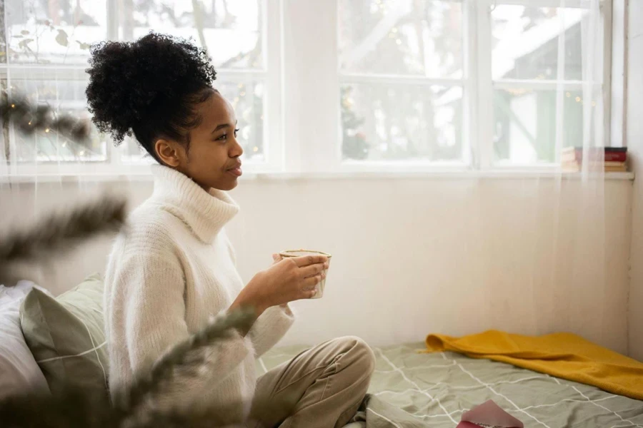 A Woman in White Knitwear Sitting on a Bed Holding a Cup