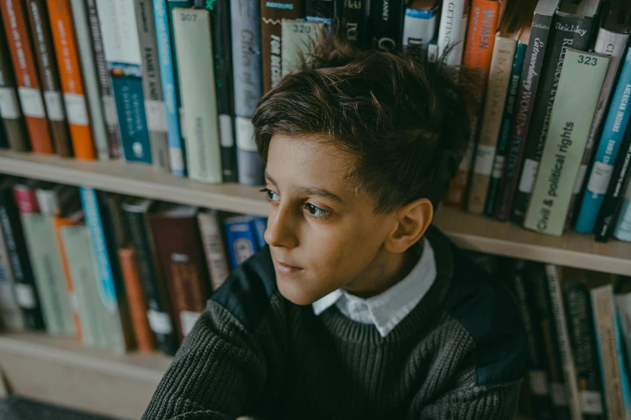 Boy Wearing a Sweater Sitting Beside Book Shelf