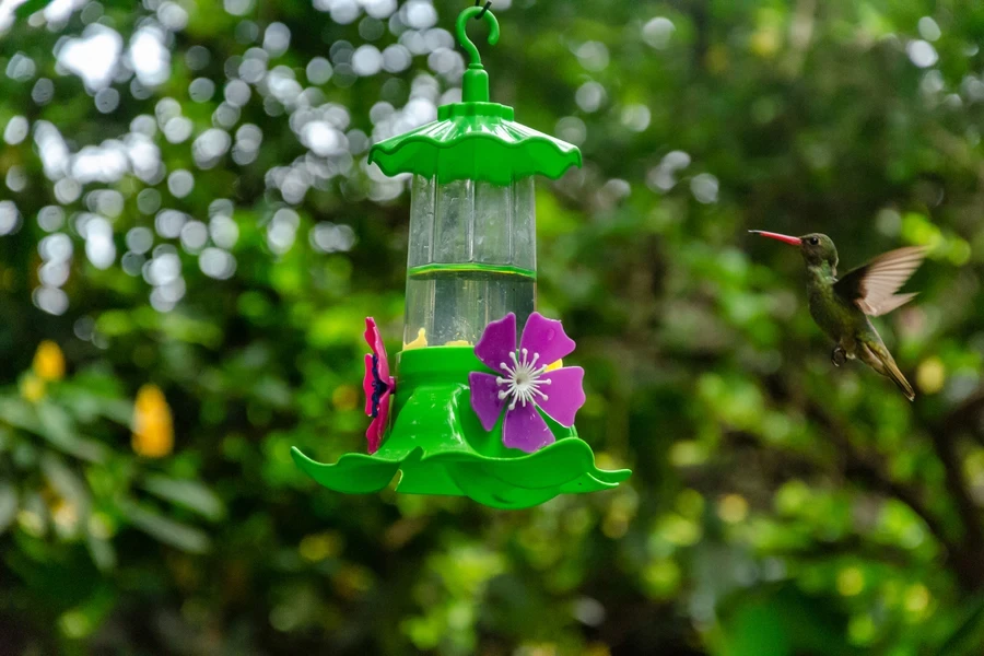 Hummingbird flying towards a green hummingbird feeder with vibrant colors