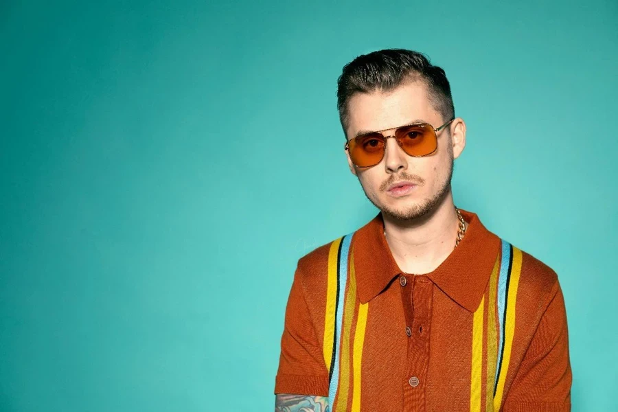 Portrait Photo of Man in Sunglasses and Brown and Yellow Polo Shirt Standing In Front of Blue Background