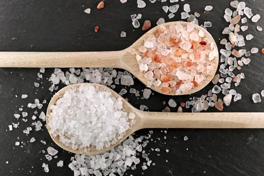 Top view of two different types of coarse salt in wooden cooking spoons on a black slate background, white sea salt and pink Himalayan rock salt