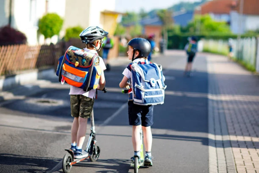 Two School Kid Boys in Safety Helmet Riding with Scooter in the City with Backpack on Sunny Day