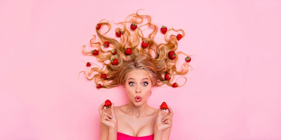 Woman with long curly wavy hair arms hands on berries wondering expression isolated pink background