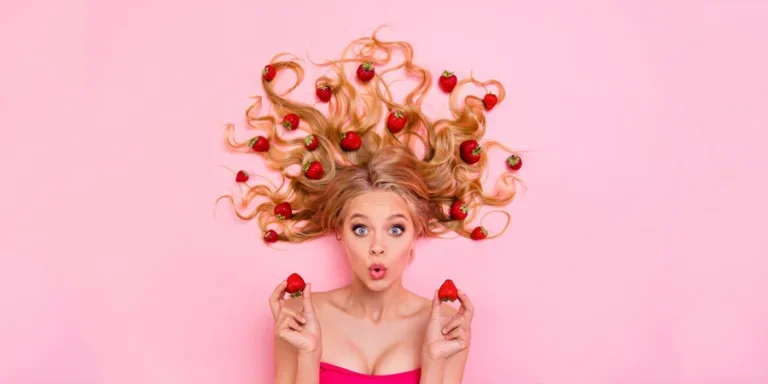 Женщина с длинными вьющимися волнистыми волосами руки на ягодах удивленное выражение изолированное на розовом фоне