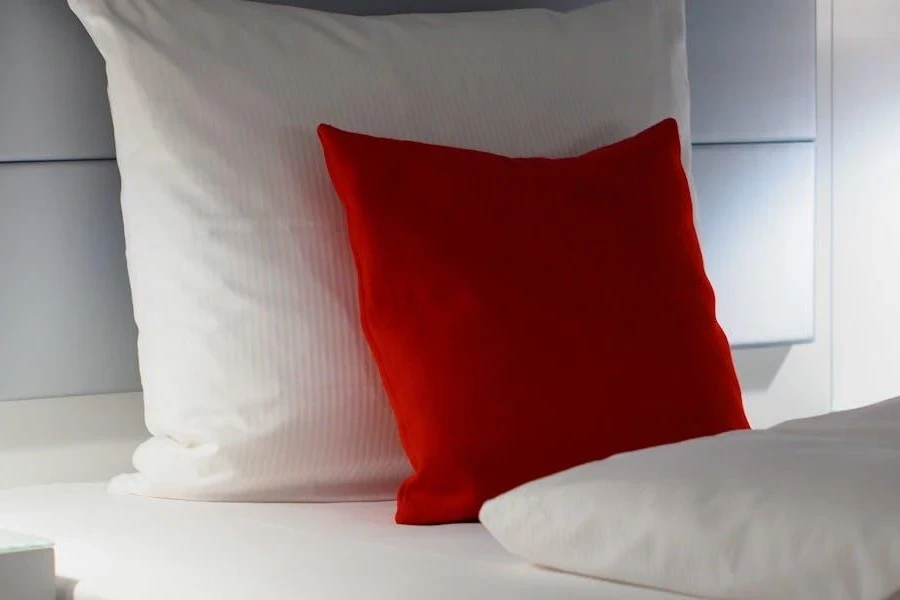 A small red cushion and big white pillow on a bed