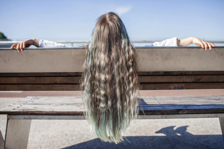 Brunette preteen girl with blue color highlights sitting on the bench on the bridge and looking at river, sunny summer day, back view, natural beauty