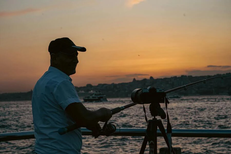 Fisherman standing beside a camera on a tripod by waterfront