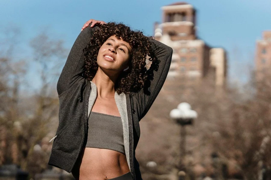 Sportive ethnic female in trendy sportswear stretching arms standing on street with trees and building on blurred background during workout