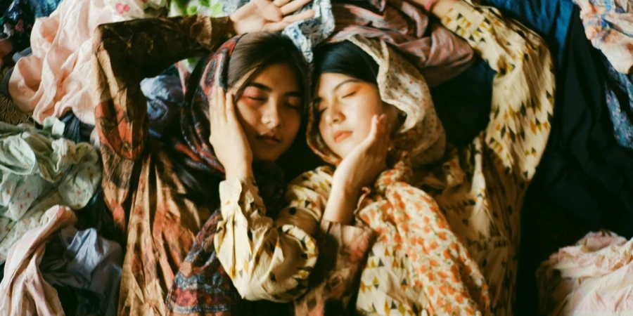 Two Women Wearing Earth-toned Printed Tops And Scarves Lying On A Pile Of Clothes