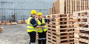 Two warehouse workers deliberate over wooden pallet stacks