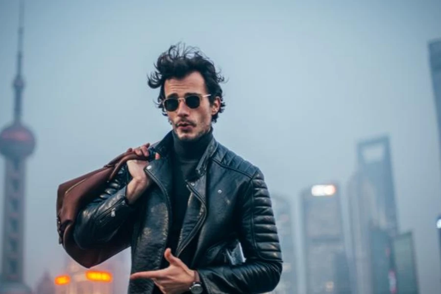 A young hipster with a leather bag standing by a fence on a foggy day