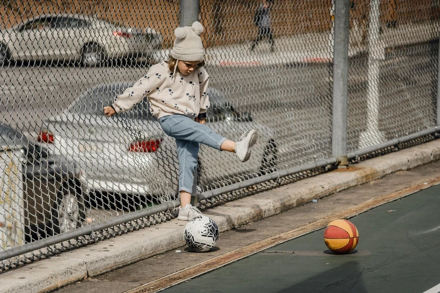 Full body of playful preschool kid in hat kicking ball while standing on sports ground near metal grid on street