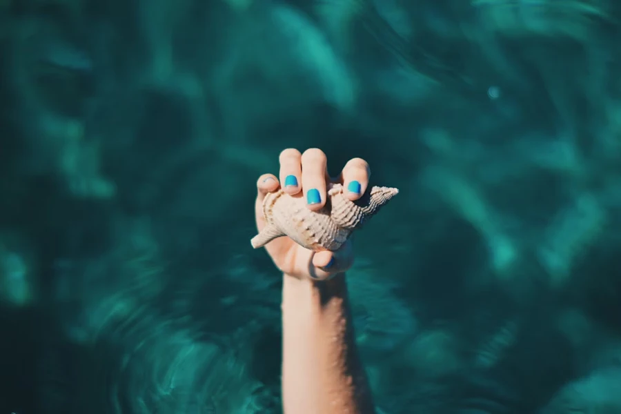 Hand with blue nail polish holding seashell in water