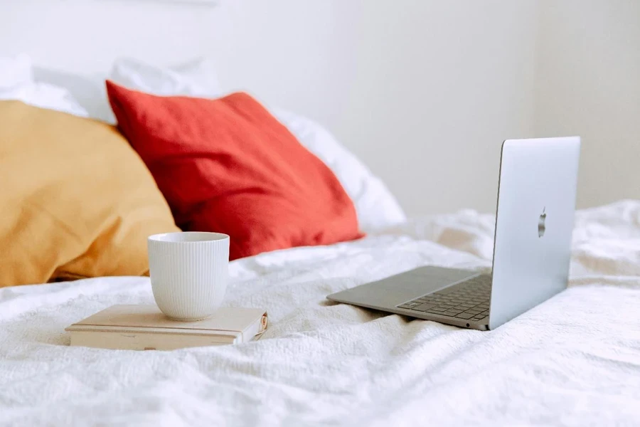 Laptop and cup of coffee on notepad near pillows on bed