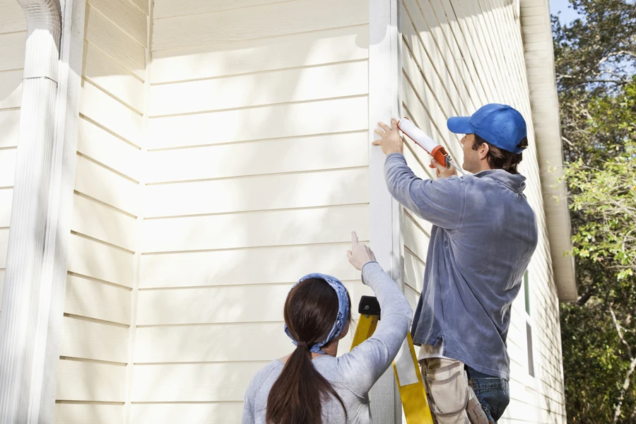 Man standing on a ladder outdoors, using a caulk gun