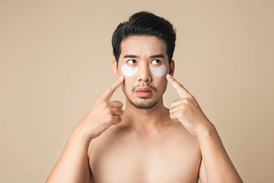 Shirtless man pointing to under eye mask he’s wearing